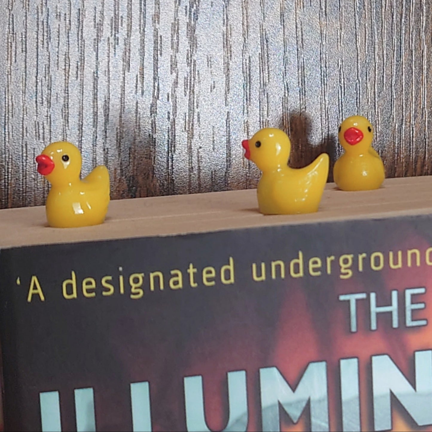 Three yellow rubber duck bookmarks on a book titled 'The Illuminatus Trilogy!' with a wooden background.