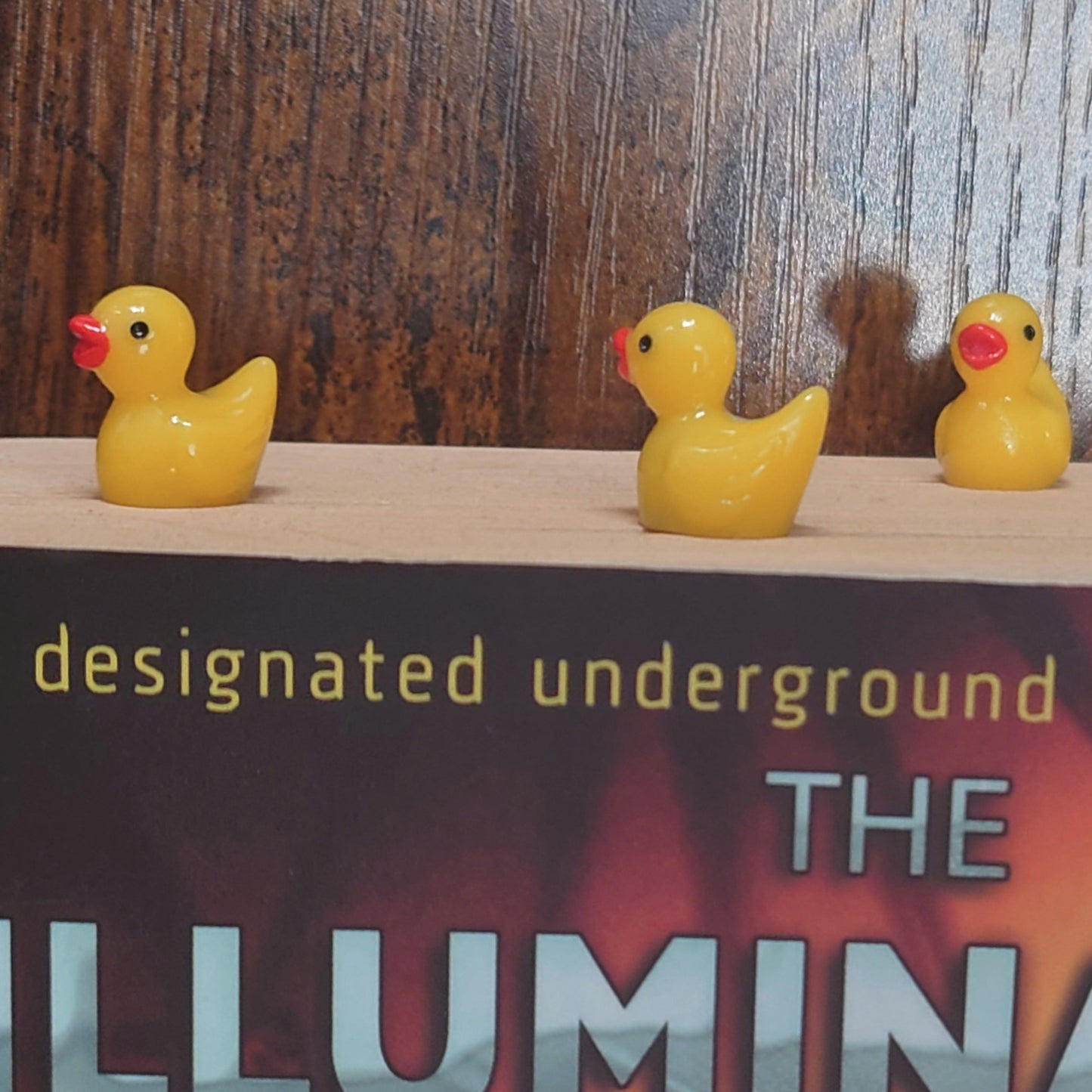 Three yellow rubber duck bookmarks on a book titled 'The Illuminatus Trilogy!' with a wooden background.
