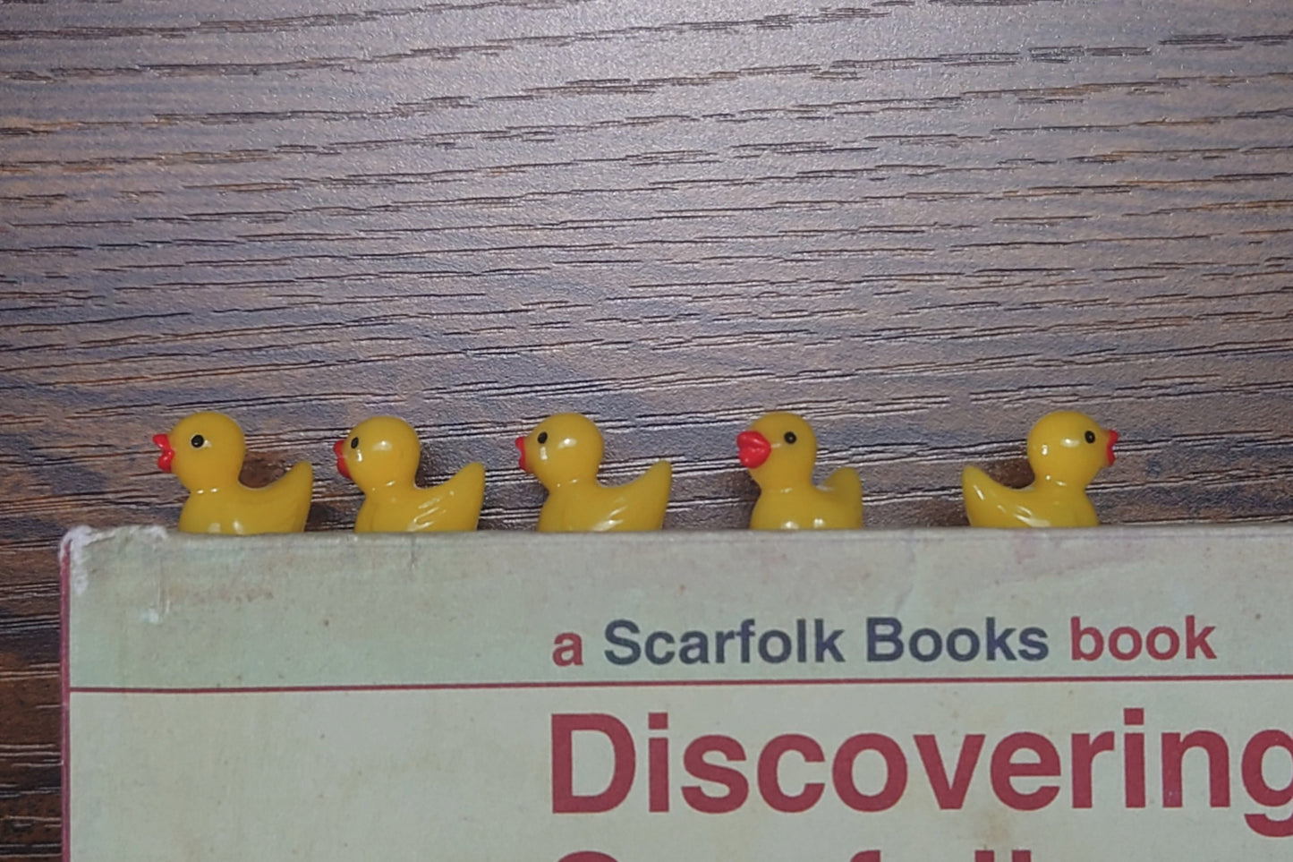 Five yellow rubber duck bookmarks on a book titled 'Discovering Scarfolk' with a wooden background.