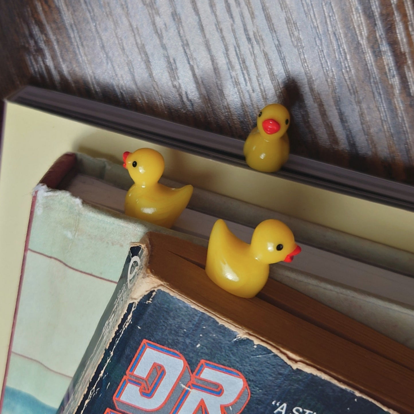 Three yellow rubber duck bookmarks in three books with a wooden background.