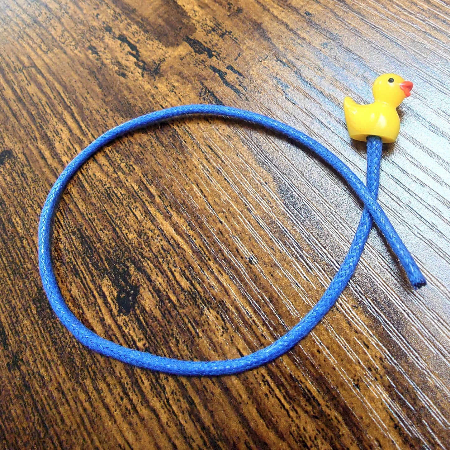 A yellow rubber duck bookmark on a wooden background with blue waxed cotton cord.