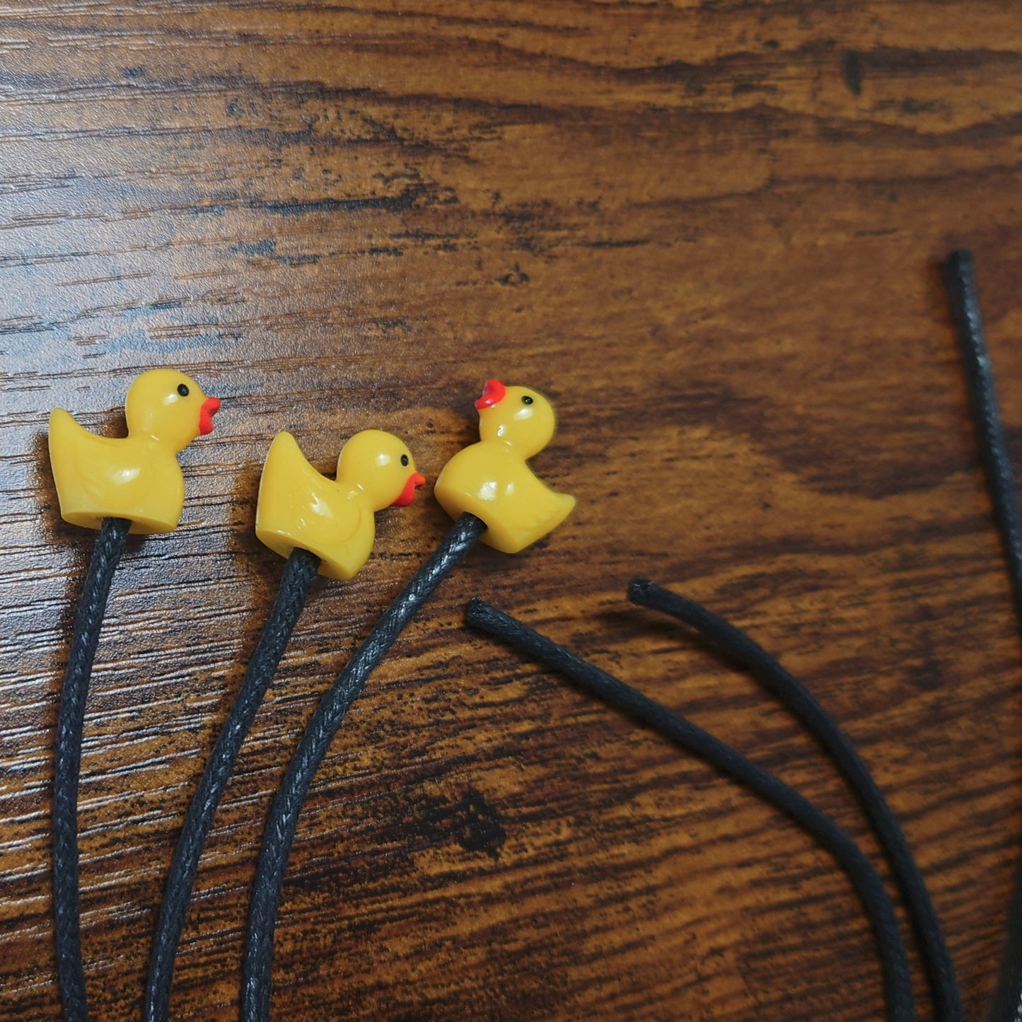 Three yellow rubber duckie bookmarks with black waxed cotton cords against a wooden background