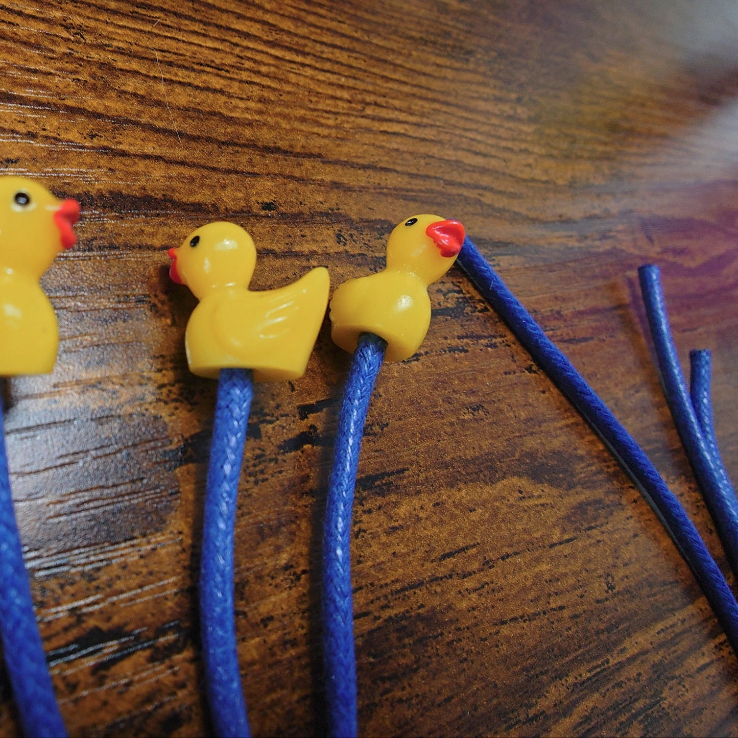 Three yellow rubber duckie bookmarks with blue waxed cotton cords against a wooden background