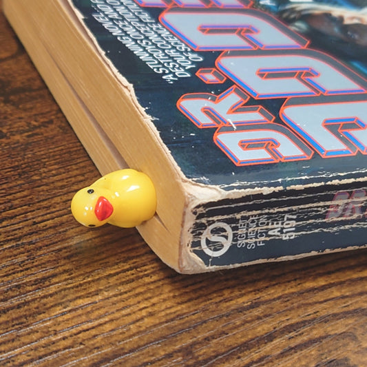 Small yellow duck bookmark with a red beak on top of a sci-fi book on a wooden surface.