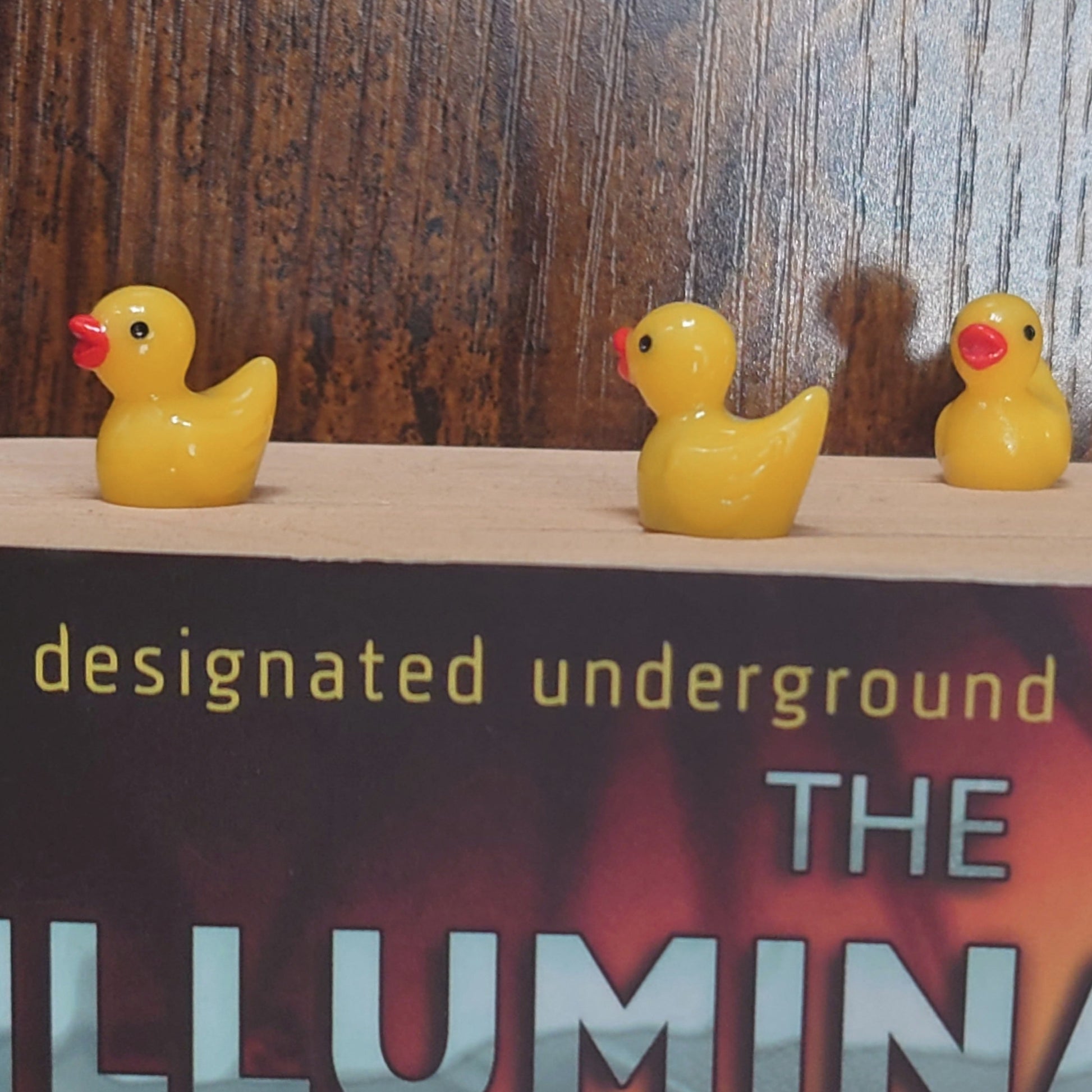 Three yellow rubber duck bookmarks on a book titled 'The Illuminatus Trilogy!' with a wooden background.